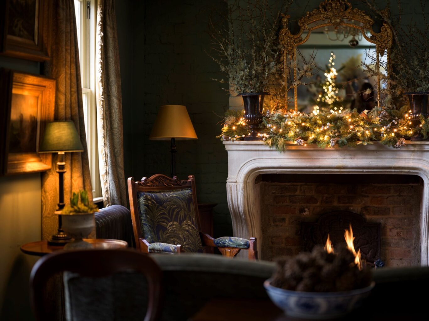 Cosy sitting room at The Rookery with a decorated fireplace, glowing garland, antique furniture, and a Christmas tree reflected in a gilded mirror